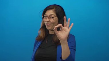 Stüdyoda el işareti gösteren ve işaret parmağını dostça gülümseyerek canlı mavi duvarda kaldıran bir kadın..
