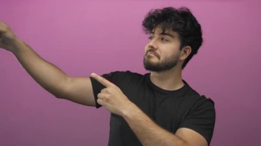 Sakallı genç adam parlak pembe bir arkaplanı işaret ediyor, yön veriyor ve özgüvenli bir ifade ve günlük siyah tişörtle odaklanıyor..