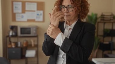 Ofiste bilek tutan, gözlüklü ve takım elbiseli esmer saçlı ve ciddi ifadeli bir kadın var..