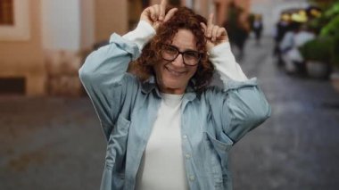 İspanyol orta yaşlı, gözlüklü kadın parmaklarıyla boynuz hareketi yapıyor. Şehrin ortasında neşeli bir ifadeyle duruyor..