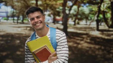 Genç adam parkta çizgili gömlek ve sırt çantası ile neşeli ifadeli defterler tutuyor parmaklarıyla zafer jestleri yapıyor..