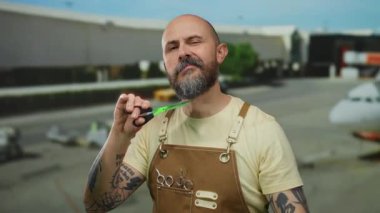 Dövmeli kel bir adam havaalanında elinde makasla poz veriyor. Terminalin dış tarafında önlük takıyor. Arka planda bulutlu bir gökyüzü ve uçak görünüyor..