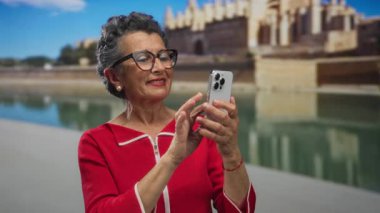Tarihi bir şehir ortamında akıllı telefon kullanan, kırmızı bir bluz ve gözlük takan, yaşlılar arasında teknolojik bir etkinlik sergileyen, gri saçlı yaşlı bir kadın..