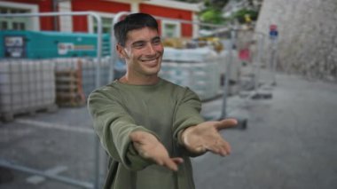 Erkek, açık hava inşaat sahasında yığılmış paletler ve malzemelerle zeytin gömleği giyerek kollarını açar; davet sıcaklığı.