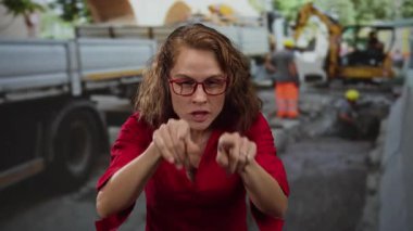 Kırmızı gömlekli kadın, arka planda işçi ve ekipman bulunan bir inşaat alanına kızgın bir şekilde bakıyor..