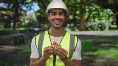Güneşli parkta waffle yerken şapkalı ve yansıtıcı yelek giyen genç İspanyol adam sırıtıyor. Keyfini çıkarın..