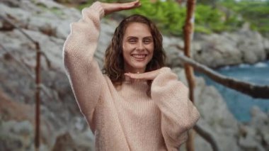 Kayalık bir sahil kenarında gülümseyen, rahat bir kazak giyen, elleriyle neşeli bir jest yapan, tasasız bir sahilin özünü yakalayan bir kadın..