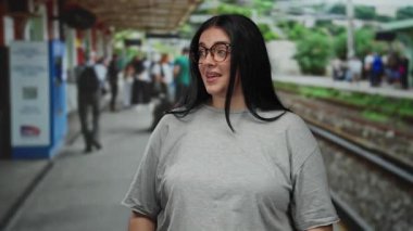 Tren istasyonunda gözlüklü genç bir kadın, düşünceli bir gülümseme ile, dış demiryolu manzarası ile çevrili ve arka planda yürüyen bulanık insanlar.