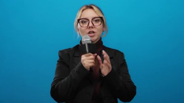 Mikrofon takan, gözlüklü, izole edilmiş mavi arka plan duvarına bakan kadın muhabir tutkuyla konuşuyor..