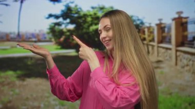 Güneşli bir parkta yeşil ağaçlar ve taş bir çitle çevrili pembe bir tişört giymiş, gülümseyen ve dışarıyı gösteren bir kadın..