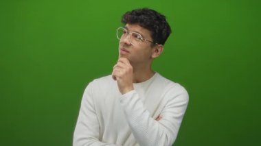 Kıvırcık saçlı gözlüklü ve eli çenesinde beyaz kazaklı bir adam stüdyoda yukarı bakıyor, düşünüyor..