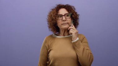 Esmer, orta yaşlı, İspanyol bir kadın, izole edilmiş mor bir duvara karşı, gözlük takıyor, düşünceli ve sıradan görünüyor..