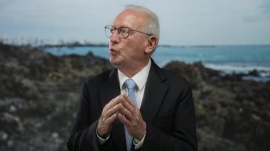 Takım elbiseli son sınıf öğrencisi, deniz kenarında özenle dikilip arka planda okyanusla birlikte profesyonel ve sakin sahil ortamının bir karışımını sergiliyor..