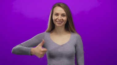 Genç bir kadın, parlak mor bir arkaplan karşısında baş parmağıyla gülümsüyor, gri bir bluz giyiyor, sade ve temiz bir ortamda mutluluk ve pozitifliği iletiyor..
