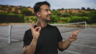 Güneşli bir tenis kortunda genç bir İspanyol, temiz bir gökyüzü altında yemyeşil ile çevrili, kendine güveni ve pozitifliğini ifade ederken iyi bir jest yapıyor..