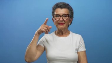 Beyaz gömlekli, kendine güveni ve mizahı olan gri saçlı ve gözlüklü yaşlı bir kadın..