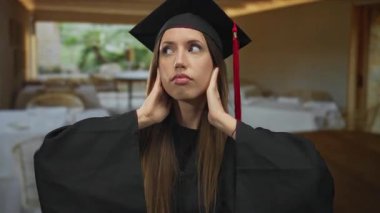 Woman in black graduation gown and mortarboard gently holds her cheeks beside tables in a cozy restaurant interior; achievement pride.