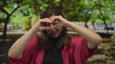 Pembe tişört giyen kadın, güneşli orman ortamında elleri ile kalp hareketi yapıyor; şefkat bağlantısı olumluluk.