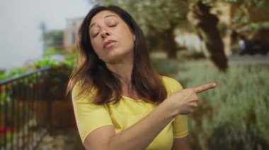 Sarı gömlekli kadın yeşil yeşilliklerin ortasında bina cephesinde parmağını sağa doğru gösteriyor; iddia makamı.