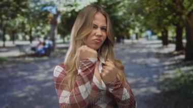 Genç, beyaz, sarışın kadın, parlak güneş ışığı altında yeşil parkta yürürken yumruklarını sıkar. Öfke, meydan okuma, saldırganlık, saldırganlık..