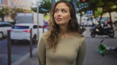 Kalabalık bir şehir sokağında gülümseyen genç İspanyol kadın arka planda trafik varken doğal ışıkta yakalanmış, rahat ve rahat bir tavır sergiliyor..