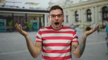 Canlı bir şehir sokak ortamında gülümseyen ve el kol hareketi yapan genç adam renkli dövmeler sergiliyor ve gözlük takıyor ve çizgili bir gömlek giyiyor..