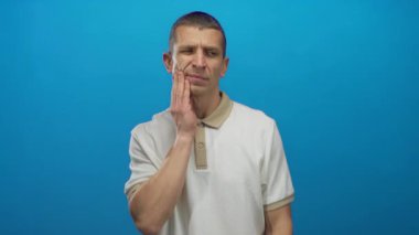 Çenesi ağrıyan bir adam mavi arkaplana karşı yanak tutarken arkasında izole bir duvar olan sıradan bir gömlek giyiyor..
