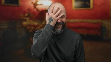 Yüzü sakallı kel bir adam kırmızı iç dekorlu zarif bir müzede dururken.