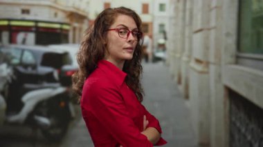 Çapraz kollu genç bir kadın hareketli bir şehir caddesinde duruyor. Kızgın ve düşünceli görünüyor. Gün boyunca dışarıda kırmızı bir gömlek ve gözlük takıyor..