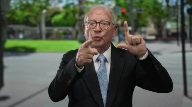 Takım elbiseli kıdemli bir adam açık havada, şehir sokaklarında, güpegündüz el kol hareketi yapıyor..