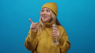 Su şişesiyle gülümseyen, sarı süveter giyen ve bereli bir kadın, izole edilmiş mavi bir arkaplanı işaret ediyor, hidrasyon ve pozitifliği gösteriyor..