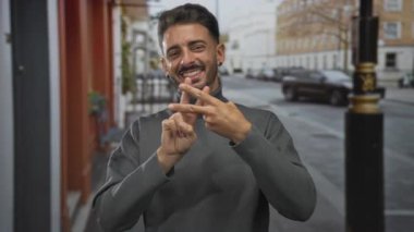 Gri balıkçı yaka giyen bir adam, gündüz vakti şehir sokaklarında parmaklarıyla hashtag sembolü yapıyor..