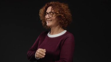 Siyah bir arkaplana karşı kollarını kavuşturarak güvenle gülümseyen kıvırcık saçları ve şık gözlükleriyle vurgulanmış zarif ve özgüvenli bir kadın..