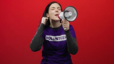 Kadın megafonla tutkuyla bağırıyor. Gönüllü gömleği ile kırmızı arka plana vuruyor. Enerjik bir gösteride vurgu yapmak için yumruğunu sıkıyor..