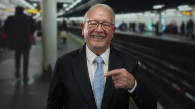 Takım elbiseli son sınıf öğrencisi başparmak kaldırıyor ve yoğun bir tren istasyonunu işaret ediyor. İçeride coşku ve pozitiflik gösteriyor..