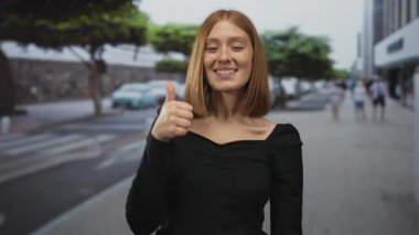 Şehir sokaklarında ağaçlar ve insanlarla gülümseyen kadın, şehir ortamında olumlu bir jest gösteriyor, açık hava kentsel ortamında pozitiflik ve kendine güven öneriyor.