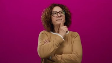 Gözlüklü kadın canlı pembe bir arka plana karşı şşşş hareketi yapıyor, kahverengi bir kazak giyiyor ve zarafet ve gizem saçıyor..