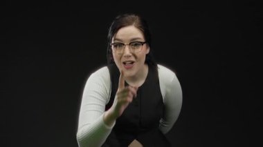 Esmer saçlı ve gözlüklü bir kadın siyah arka plana karşı sessiz bir jest yapıyor. Gizlilik ve sessizliği ifade ediyor.