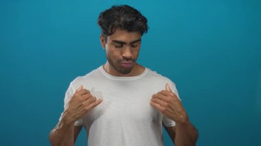 Genç İspanyol adam, mavi stüdyo ortamında beyaz gömleği göğsünün üzerinde iki eliyle ayarlar..