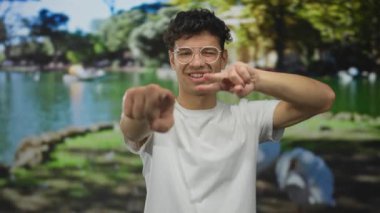 Göl kenarındaki yeşil orman parkında gözlüklü bir adam kamerayı işaret ediyor..