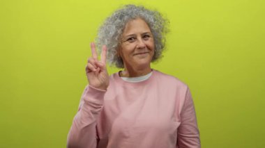 Gri saçlı, gülümseyen ve hayat dolu sarı bir arka plana barış işareti koyan pembe kazaklı, mutluluk ve pozitiflik taşıyan bir kadın..