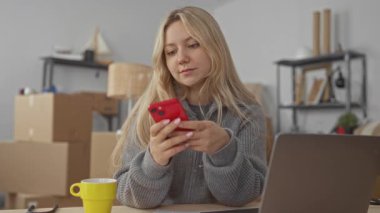 Yeni apartman dairesinde çalışan bir kadın dizüstü bilgisayar ve telefonla etrafı taşınan kutular ve ev eşyalarıyla çevrili bir şekilde şık oturma odasında rahat bir ortam yaratıyor..