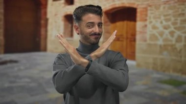 Genç İspanyol adam, eski kentteki kaldırım taşı caddesindeki yıpranmış taş binanın önünde kollarını kavuşturuyor..