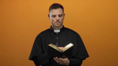 Genç rahip parlak turuncu arka planda sürpriz bir şekilde İncil okuyor, dini bağlılık ve beklenmedik keşifler sergiliyor..