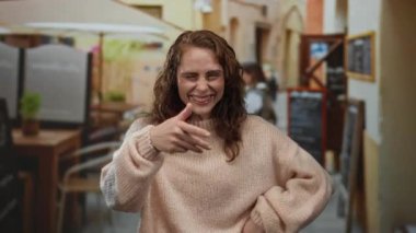 Şehir caddesinde bir restoranın terasında gülen bir kadın, rahat bir kazak giyiyor, arka planda bulanık masalar ve menü tahtaları var..