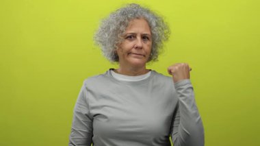 Kıvırcık gri saçlı bir kadın omuzlarının üstünden gri bir gömlek giyerek parlak sarı bir arkaplanı işaret ederek kendine güvenini ve soruşturmasını ifade ediyor.