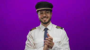 Üniformalı genç adam canlı mor bir arka plana karşı alkışlıyor, kendine güvenen, sakallı ve şapkalı, profesyonel ve enerjik bir tavır sergiliyor..