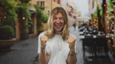 Sarışın, uzun saçlı bir adam kaldırım taşı caddesindeki açık büfe masalarının önünde yumruklarını sıkıyor. Zafer..