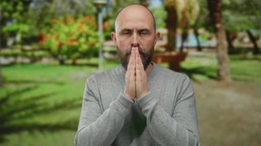 Huzurlu parkta meditasyon yapan bir adam. Dua eden elleri ve güneş ışığı altındaki sakin ifadesiyle. Gri bir süveter giyiyor.