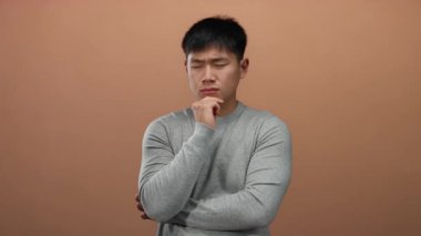 Gri gömlekli genç adam kahverengi bir arka plan üzerinde düşünceli bir ifade ile, bir stüdyo ortamında tefekkür ve içgözlem örneği..